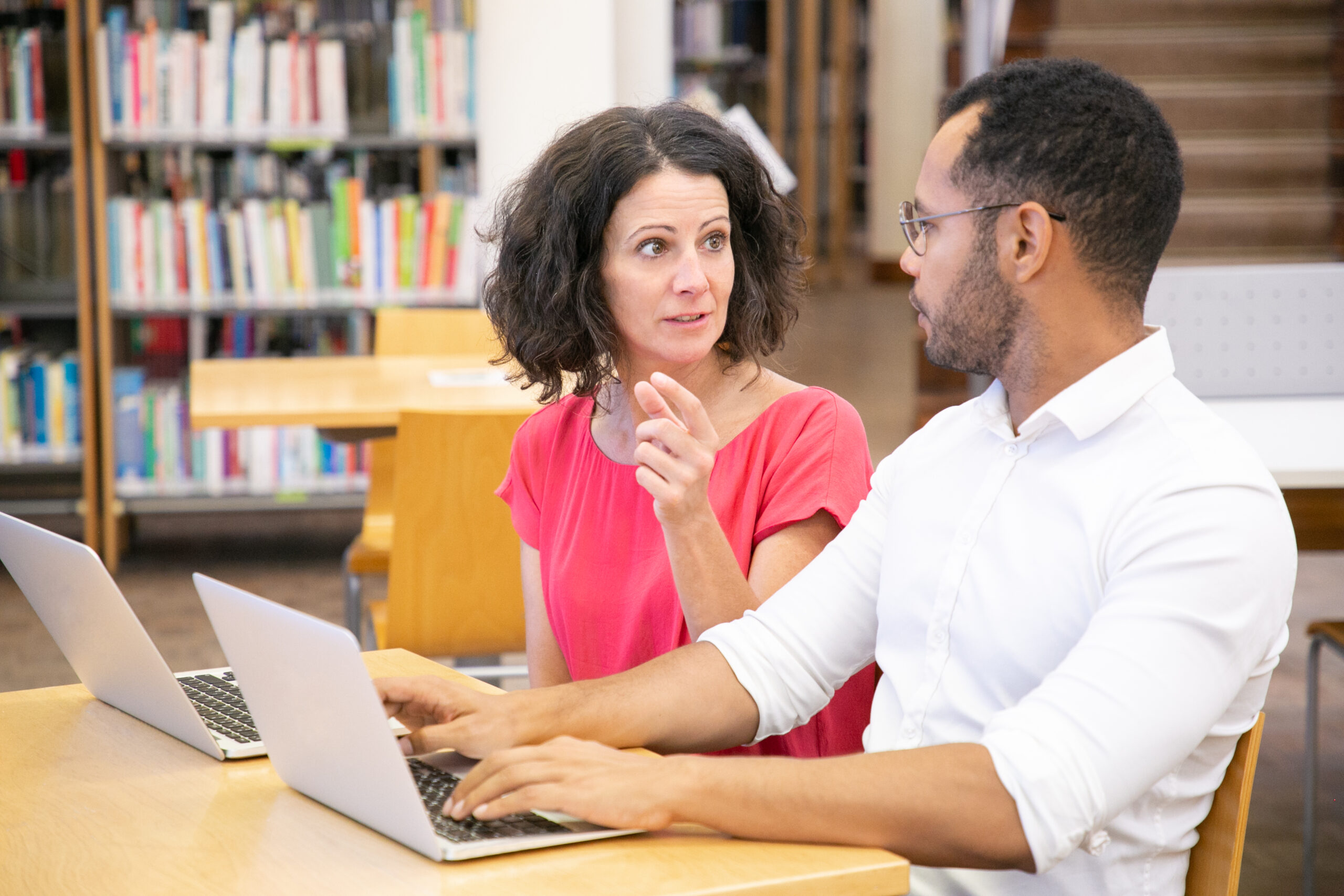 Excited adult students sharing ideas for project in library. Man and woman in casual sitting at desk, using laptops, gesturing and talking. Brainstorming concept